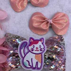 May include: Three pink and iridescent hair bows with a white cat applique. The cat is embroidered with purple and orange details.
