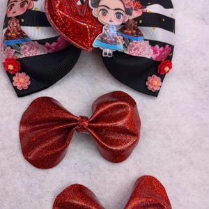 May include: Three red glitter hair bows. The largest bow features a black and white striped ribbon with a red heart and a cartoon image of Frida Kahlo.