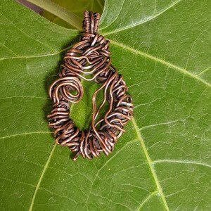 May include: A copper wire tree of life pendant, with a delicate, intricate design, is displayed on a green leaf.