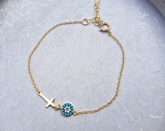 May include: A gold-tone chain bracelet featuring a small, plain cross charm and a circular evil eye charm adorned with tiny turquoise and white stones. The bracelet includes a delicate link chain with a circular spring ring clasp and an extender.