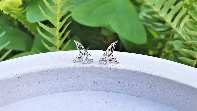 May include: A pair of silver earrings with a clear crystal in the center of each earring. The earrings are shaped like wings.