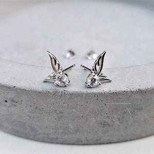 May include: A pair of silver earrings with a small clear crystal and a wing design.
