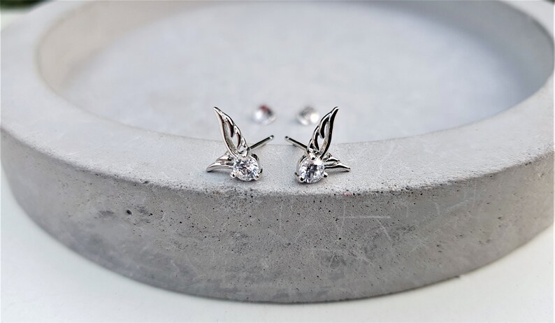 May include: A pair of silver earrings with a small clear crystal and a wing design.