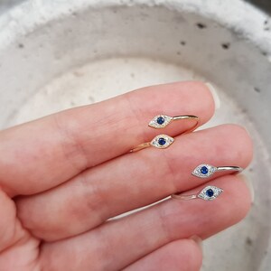 May include: Two gold and silver rings with a blue evil eye design. The rings are on a finger.