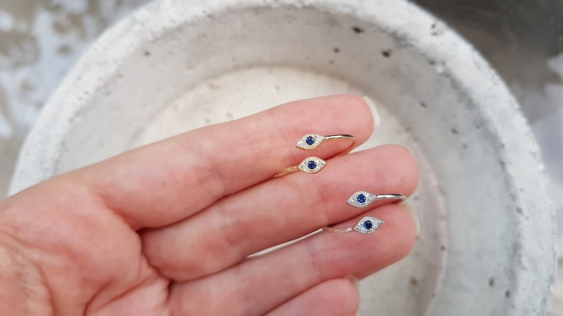 May include: Two gold and silver rings with a blue evil eye design. The rings are on a finger.