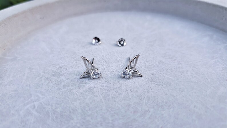 May include: A pair of silver earrings with a clear crystal in the center of each earring. The earrings are shaped like wings.