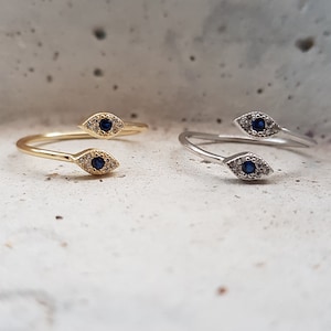 May include: Two gold and silver rings with a blue evil eye design. The rings are on a white textured surface.