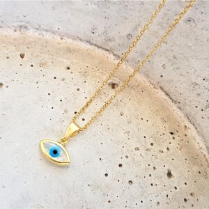 May include: A gold chain necklace with a blue evil eye pendant. The pendant is oval shaped and has a white background.