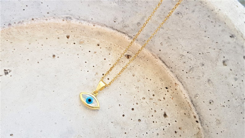 May include: A gold chain necklace with a blue evil eye pendant. The pendant is oval shaped and has a white background.