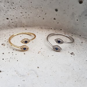 May include: Two adjustable rings with a double evil eye design. One ring is gold and the other is silver. Each eye has a blue center and is surrounded by small white stones.