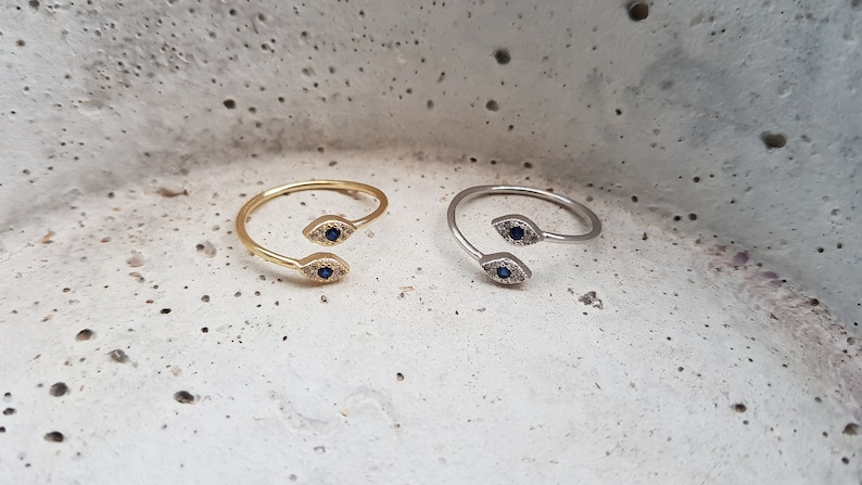 May include: Two adjustable rings with a double evil eye design. One ring is gold and the other is silver. Each eye has a blue center and is surrounded by small white stones.