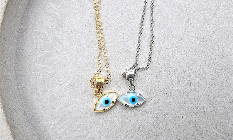 May include: Two necklaces with evil eye pendants. One necklace has a gold chain and the other has a silver chain. Both pendants are white with a blue eye.