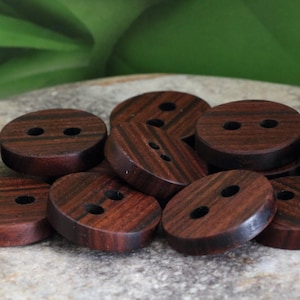 Puede incluir: Un grupo de 12 botones de madera marrón con dos agujeros cada uno. Los botones están dispuestos en una pila sobre una superficie marrón claro.