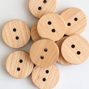 Puede incluir: Un grupo de diez botones de madera, cada uno con dos agujeros. Los botones son de color marrón claro y tienen una veta de madera natural.