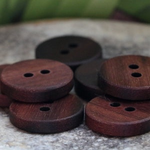 Puede incluir: Un conjunto de botones de madera marrón oscuro, cada uno con dos agujeros. Los botones son redondos y tienen un acabado liso. Algunos están apilados, otros dispersos sobre una superficie de piedra. Los botones varían ligeramente en color.