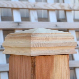 May include: A wooden post cap with a square base and a decorative, multi-layered, pyramid-shaped top. The cap is made of unfinished wood and has a natural wood grain.