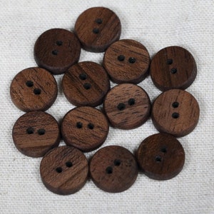 Könnte beinhalten: Eine Gruppe von 13 braunen Holzknöpfen mit jeweils zwei Löchern. Die Knöpfe sind in einem Kreis auf einem weißen Stoffhintergrund angeordnet.