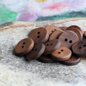 Puede incluir: Un montón de botones de madera marrón con dos agujeros cada uno. Los botones están dispuestos en un patrón aleatorio y todos son del mismo tamaño.