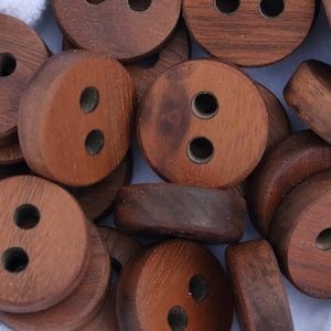 Puede incluir: Un montón de botones de madera marrón con dos agujeros cada uno. Los botones están dispuestos en un patrón aleatorio sobre una superficie blanca.