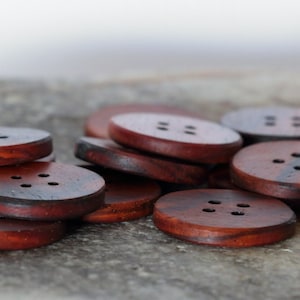 May include: A pile of dark brown wooden buttons with four holes each. The buttons are stacked on top of each other and have a natural wood grain.