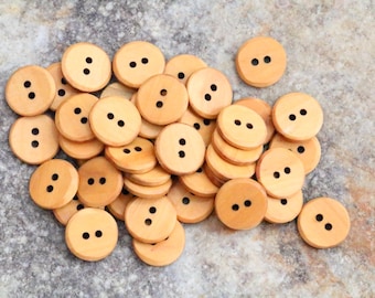 May include: A collection of light brown wooden buttons with two holes each. The buttons are round and have a natural wood grain. They are scattered on a textured surface, suitable for sewing and craft projects.