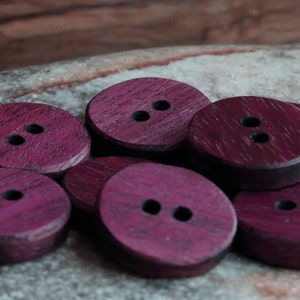 Puede incluir: Un grupo de ocho botones de madera de color morado, cada uno con dos agujeros. Los botones son redondos y tienen un borde ligeramente elevado.