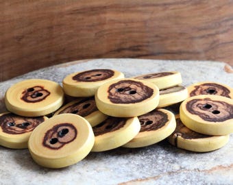 May include: A pile of wooden buttons with a natural wood grain and brown markings. The buttons are round and have two holes.