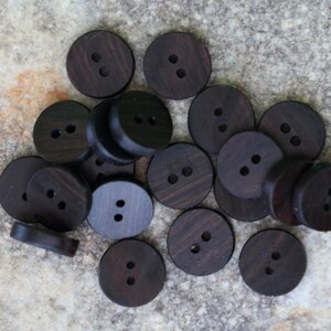 May include: A selection of dark brown, circular wooden buttons, each with two holes. The buttons are arranged on a textured, light brown surface. These buttons are ideal for sewing and craft projects.