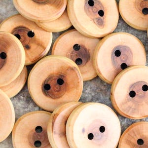 May include: A selection of wooden buttons, each with two black holes. The buttons are circular, displaying a light brown hue and visible wood grain. The buttons are scattered on a light grey surface.