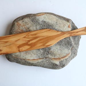 May include: A wooden spatula with a natural wood grain finish. The spatula is resting on a gray rock.
