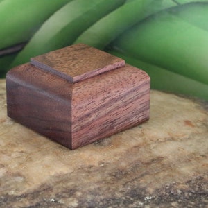 May include: A small, square wooden box with a dark brown lid and a light brown base. The box is made of walnut wood and has a natural wood finish.