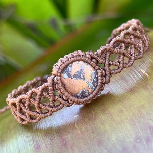 Peut inclure: Un bracelet en macramé marron doté d'une pierre circulaire centrale à la surface tachetée de cuivre et de gris foncé. Le bracelet est composé d'un cordon marron noué de manière complexe, créant un motif en treillis entourant la pierre.