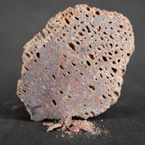 May include: A close-up of a piece of red and gray porous rock with a rough texture. The rock has many small holes and is broken into several pieces.