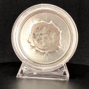 May include: A silver coin with a textured surface is displayed in a clear plastic coin holder with a stand.