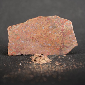 May include: A close-up of a red and brown stone with a mottled pattern. The stone is flat and has a rough texture. There are small pieces of the stone scattered around it.