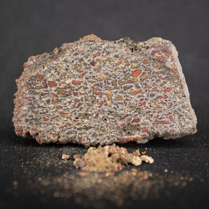 May include: A close-up of a rough, gray rock with red and orange veins. The rock has a unique, layered pattern and is surrounded by small, loose pieces of the same material.