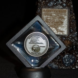 May include: A silver coin commemorating the "First Nuclear Test" of July 16, 1945, encased in a clear display case. The coin features a depiction of a helmet and a piece of trinitite. A decorative, textured object is in the background.