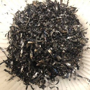 May include: A close-up shot of a pile of metallic shavings, appearing dark gray and silver. The shavings are curled and ribbon-like, with a reflective sheen. The texture is rough and irregular, suggesting a manufacturing process.