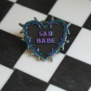 May include: A black heart-shaped pin with a barbed wire border. The pin has a rainbow iridescent finish and says "SAD BABE" in purple letters.