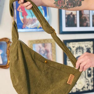 May include: Olive green corduroy shoulder bag with a curved design. Features a long strap, a small pocket, and a leather tag with the text "MOXXIE". The bag is held up by a person.