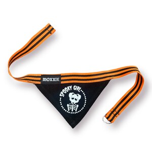 May include: Black dog bandana with orange and black striped trim. The bandana has a white graphic of a dog skeleton with the text "Spooky Girl" around it. The bandana has a metal ring for attaching to a collar.