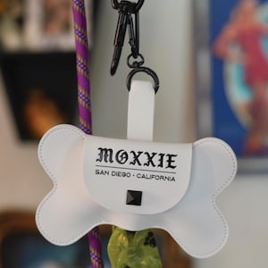 May include: White bone-shaped dog waste bag dispenser with the text "MOXXIE SAN DIEGO - CALIFORNIA" and a black stud. The dispenser is attached to a purple and gold rope.
