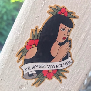 Könnte beinhalten: Ein Sticker mit einer Schwarzweißillustration einer Frau, die ihre Hände zum Gebet faltet. Sie trägt ein schwarzes, ärmelloses Oberteil und hat rote Blumen im Haar. Der Sticker hat ein goldenes Banner mit dem Text "PRAYER WARRIOR" in Schwarz.