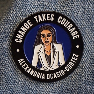 May include: A black and white enamel pin with a blue background. The pin features a cartoon portrait of Alexandria Ocasio-Cortez and the text "Change Takes Courage Alexandria Ocasio-Cortez".