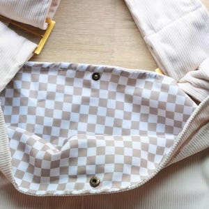 Op de afbeelding: Beige corduroy tas met een geruite binnenkant. De tas heeft een lange riem en een goudkleurige gesp. De binnenvoering heeft een bruin-wit geruit patroon. De tas sluit met een drukknop.