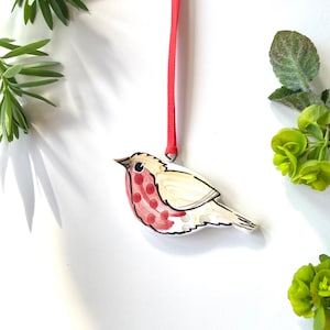 May include: A wooden bird decoration with a red hanging ribbon. The bird features a cream body, a red breast with white spots, and black outlines. Green leaves and foliage surround the ornament.