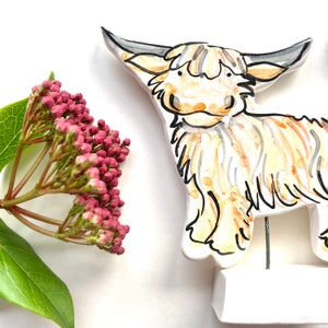 May include: A ceramic Highland cow ornament with a white base, hand-painted in tan, cream, and black. The cow has large horns and a shaggy coat. A sprig of pink flowers and green leaves is in the background.