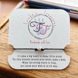 Puede incluir: Una pulsera de cordón negro con un pequeño dije de estrella plateada, montada en una tarjeta de presentación blanca. La tarjeta muestra el texto Trisha's Treasures, Handmade with love, y un poema sobre atar la pulsera a la muñeca.