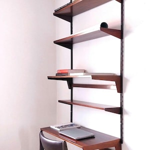 May include: A dark wood wall-mounted shelving unit with a desk and chair. The shelves hold books and a laptop. The unit is attached to a white wall. The desk and shelves are made of dark wood.