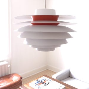 May include: A multi-tiered pendant light fixture featuring a series of stacked, circular white shades with a single vibrant orange accent ring near the top, suspended from the ceiling by a thin, straight white cord.